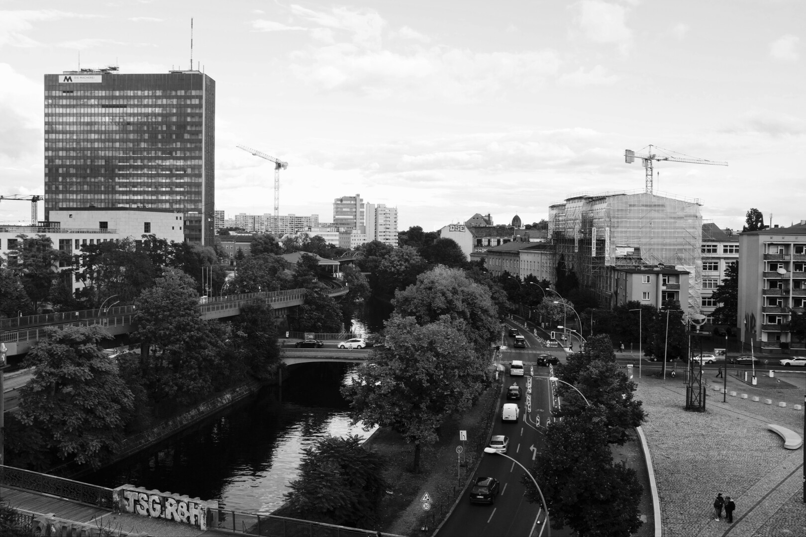 A photo taken in Berlin-Kreuzberg, showing a street with some tiny cars, a river and some typical buildings.
